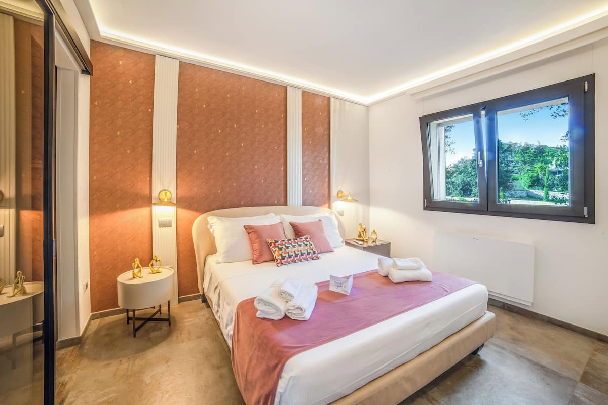 A modern hotel room with a large bed, bedside tables, a window with a view, and a mirror.