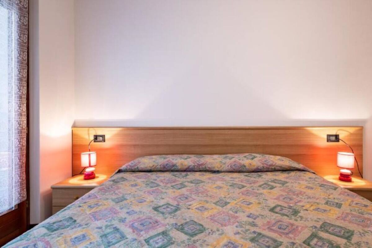 A bedroom with a wooden headboard, two bedside lamps, a patterned bedspread, and a window with a curtain.