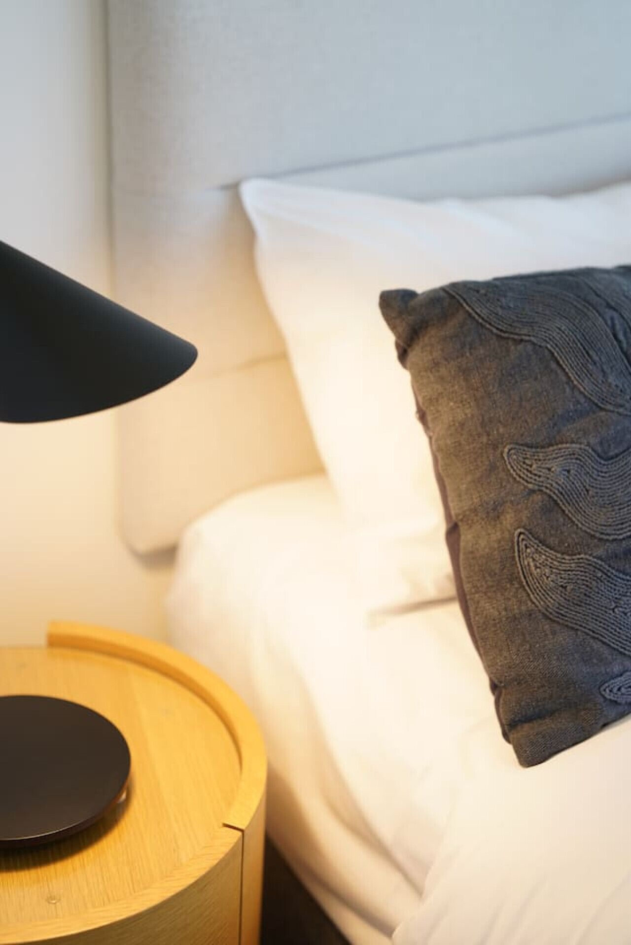 Een net opgemaakt bed met witte lakens en een donkergrijze kussens. Er staat een rond houten nachtkastje met een zwarte lamp erop.