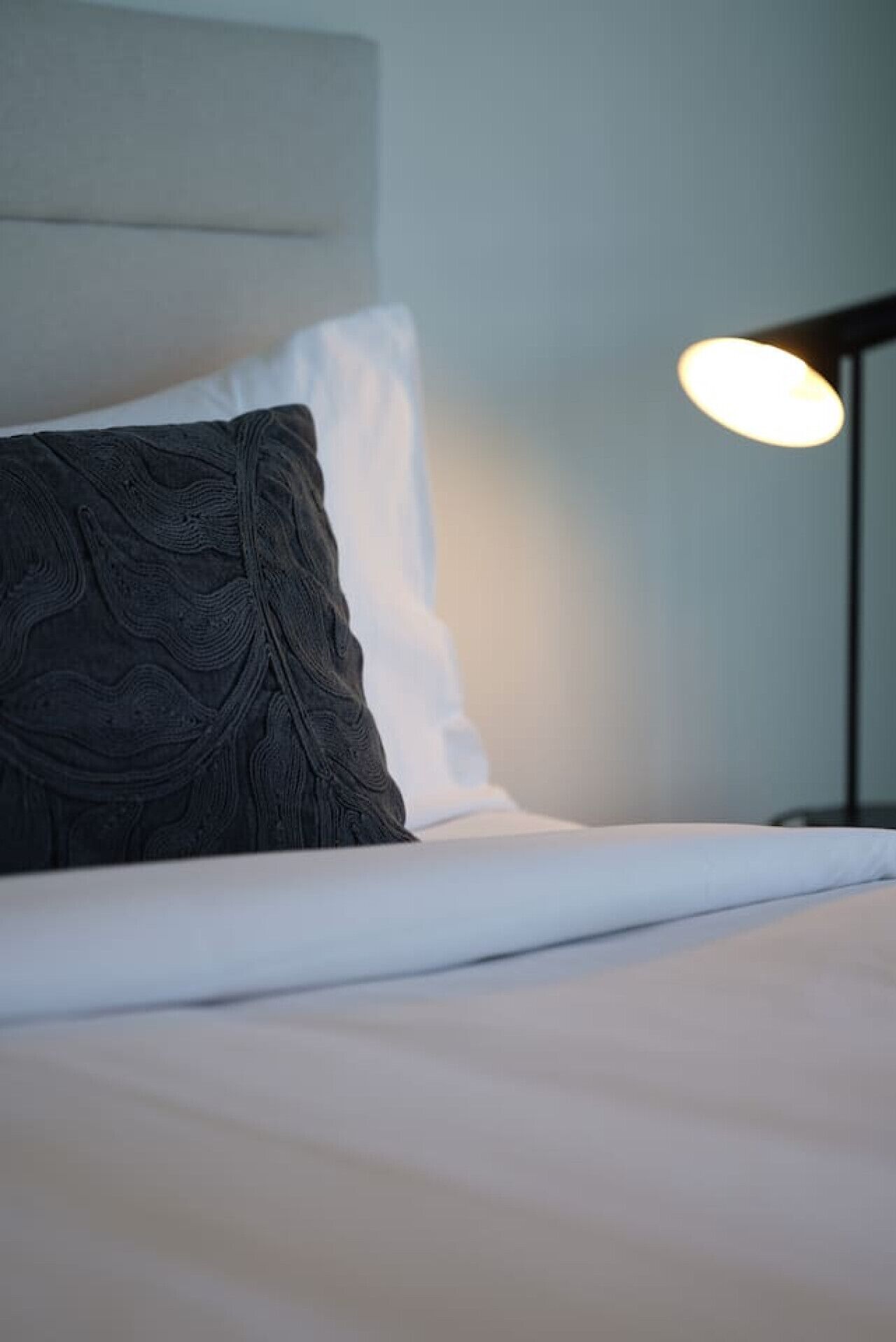 Ein ordentlich bezogenes Bett mit einem dunkel gemusterten Kopfkissen und einem weißen Kissen, einer Nachttischlampe und einer schlichten Wand im Hintergrund.