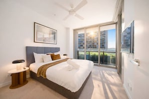 A modern bedroom with a large bed, a ceiling fan, a bedside table with a lamp, and a view of a cityscape through a sliding glass door. - Modern City 1-Bedroom | Prime CBD Location (Brisbane City)