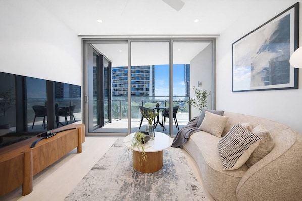 A modern living room with a flat-screen TV, a sofa, a coffee table, and a balcony with a view of buildings. - Modern City 1-Bedroom | Prime CBD Location (Brisbane City)