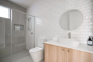 Een moderne badkamer met witte metro tegels, een douche met glazen wand, een wit toilet, een houten wastafelmeubel met een grote wasbak en een ronde spiegel.