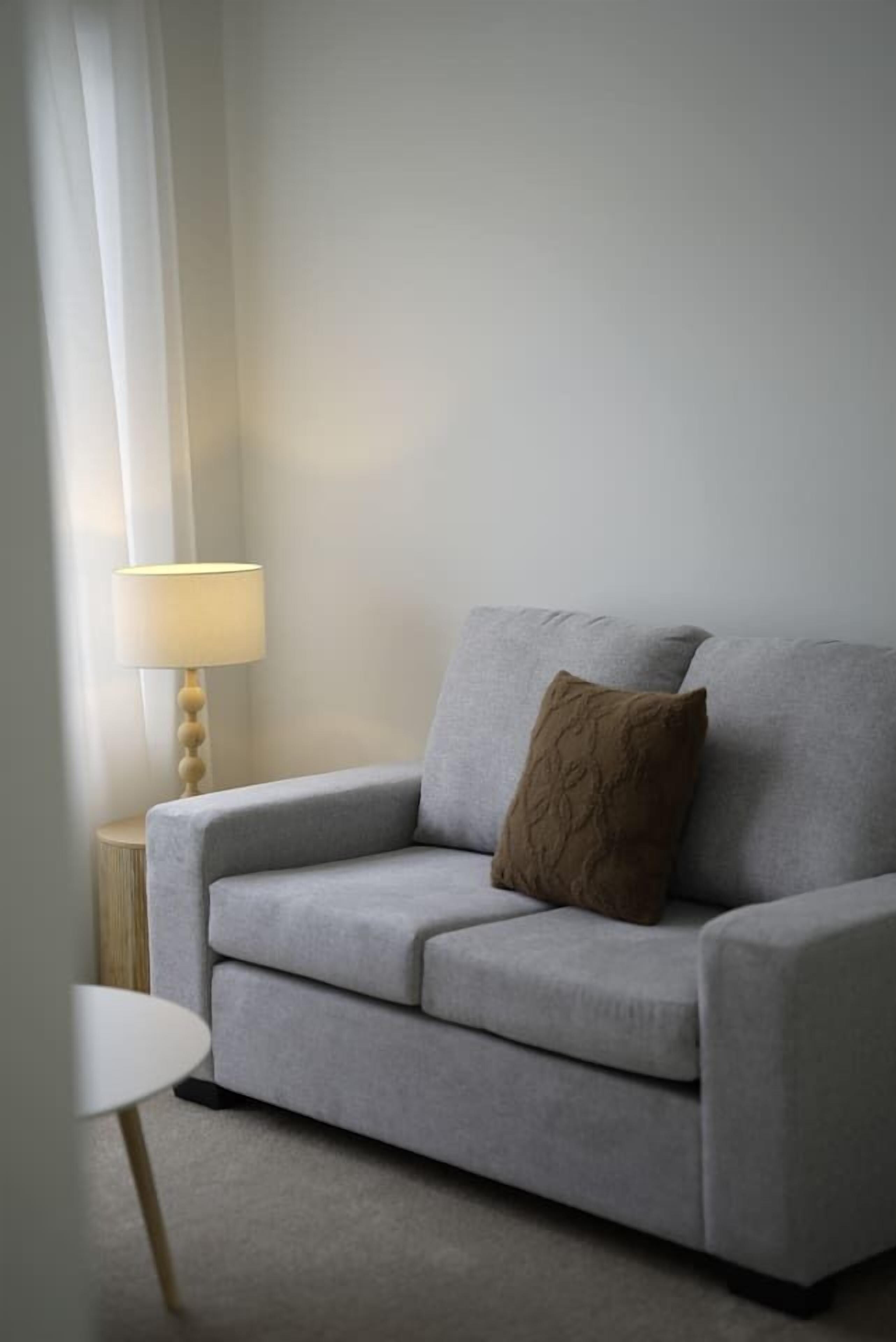 A grey sofa with a brown cushion in a room with a lamp and a small table.
