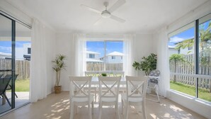 A bright, sunlit room with a ceiling fan, a dining table with white chairs, and large windows offering a view of the backyard and neighboring houses. - 3 Lounges 5 Beds w/ Ducted A/C Modern & Convenient (Rochedale)