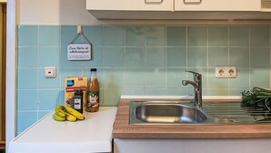 Una cocina con encimera blanca, pileta de acero inoxidable y salpicadero de azulejos azul claro. Hay plátanos, una botella de sirope y un tarro de mantequilla de maní sobre la encimera.