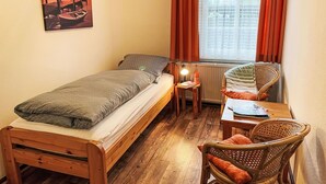 Una habitación pequeña y acogedora con una cama individual, una silla, una mesa y un ventanal con cortinas.