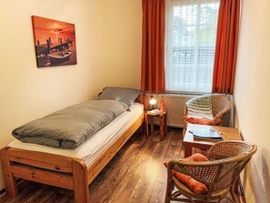 Ein kleines, gemütliches Zimmer mit einem Bett, einem Sessel, einem Tisch und einem Fenster mit Vorhängen.