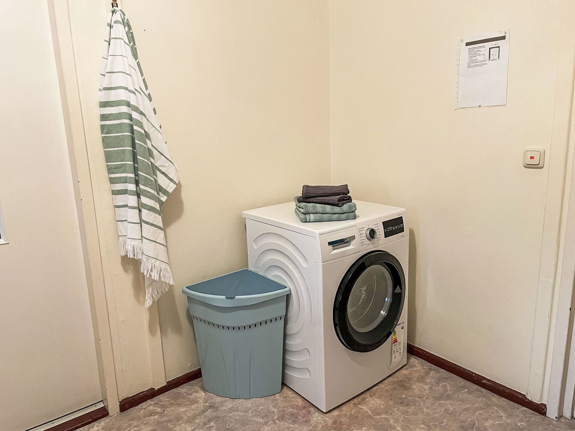 A laundry room with a washing machine, a trash bin, and a towel hanging on the wall.