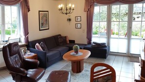 En stue med en mørk lædersofa, et træskab med glaslåger, et spisebord med en stol og store vinduer med gardiner.