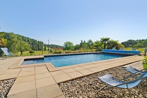 Une piscine entourée d’une terrasse carrelée, de chaises longues et offrant une vue sur une colline verdoyante.