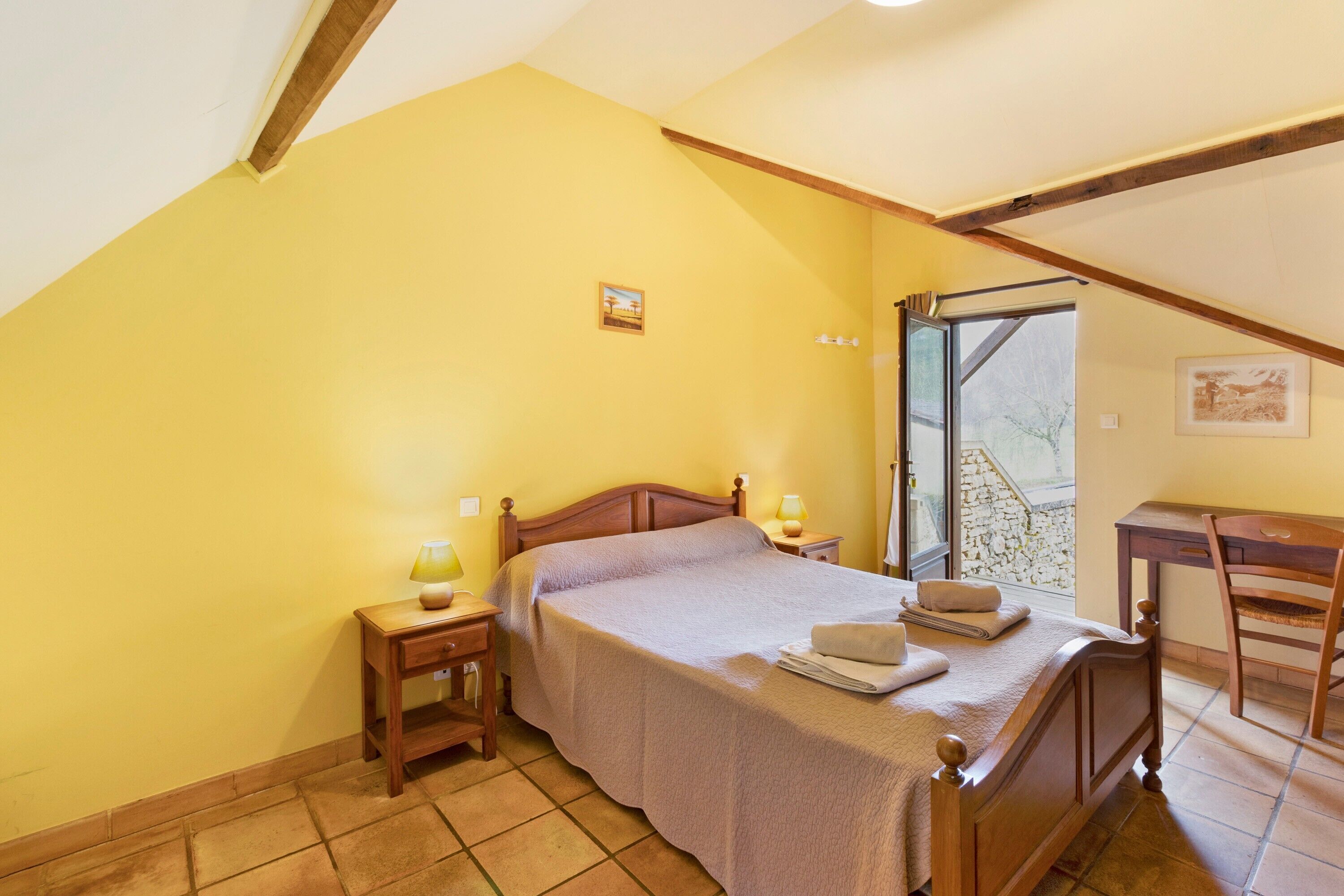 A bedroom with a wooden bed, bedside tables with lamps, a chair, a window with a view, and a small picture on the wall.