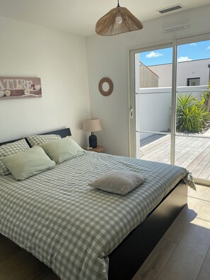 A bedroom with a checkered bedspread, a bedside lamp, a wall-mounted mirror, and a view of the outdoors through a sliding glass door. - Family home (Saint-Pierre-d'Oléron)