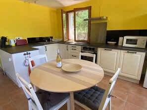 Une cuisine compacte avec des meubles blancs, une table ronde entourée de chaises rembourrées, une bouteille de vin et un four à micro-ondes.