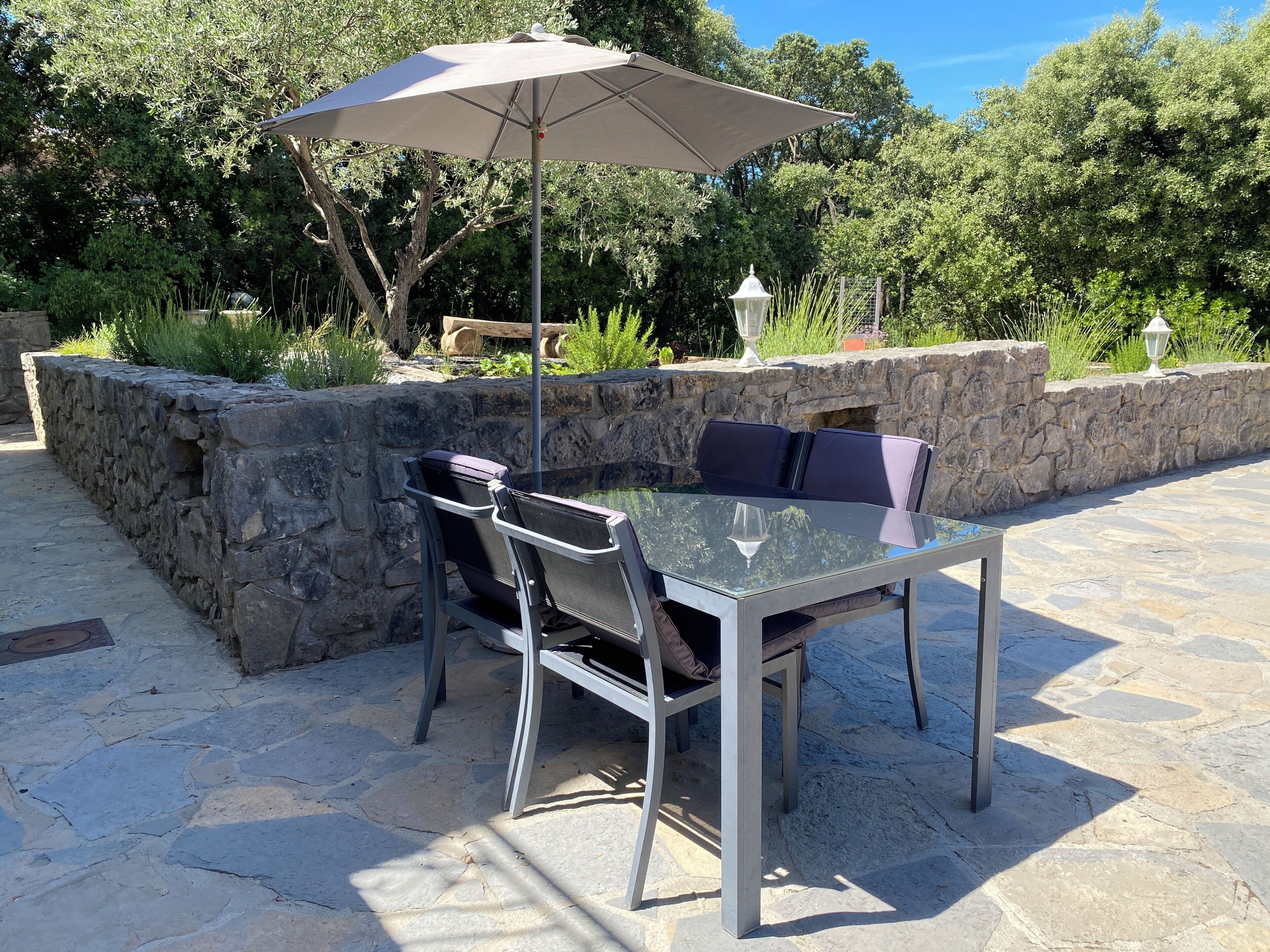 Un patio de piedra con una mesa y sillas de vidrio, una sombrilla y un muro de piedra.