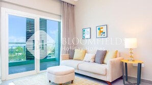 A modern living room with a sofa, ottoman, and a small table with a lamp. There are two framed pictures on the wall and a large window with a view of greenery. - Bloomfields Deluxe 1Br Leonardo (Abu Dhabi)