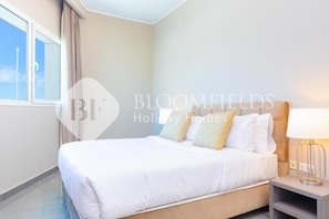 A hotel room with a bed, a nightstand, a lamp, and a window with curtains. - Bloomfields Deluxe 1Br Leonardo (Abu Dhabi)