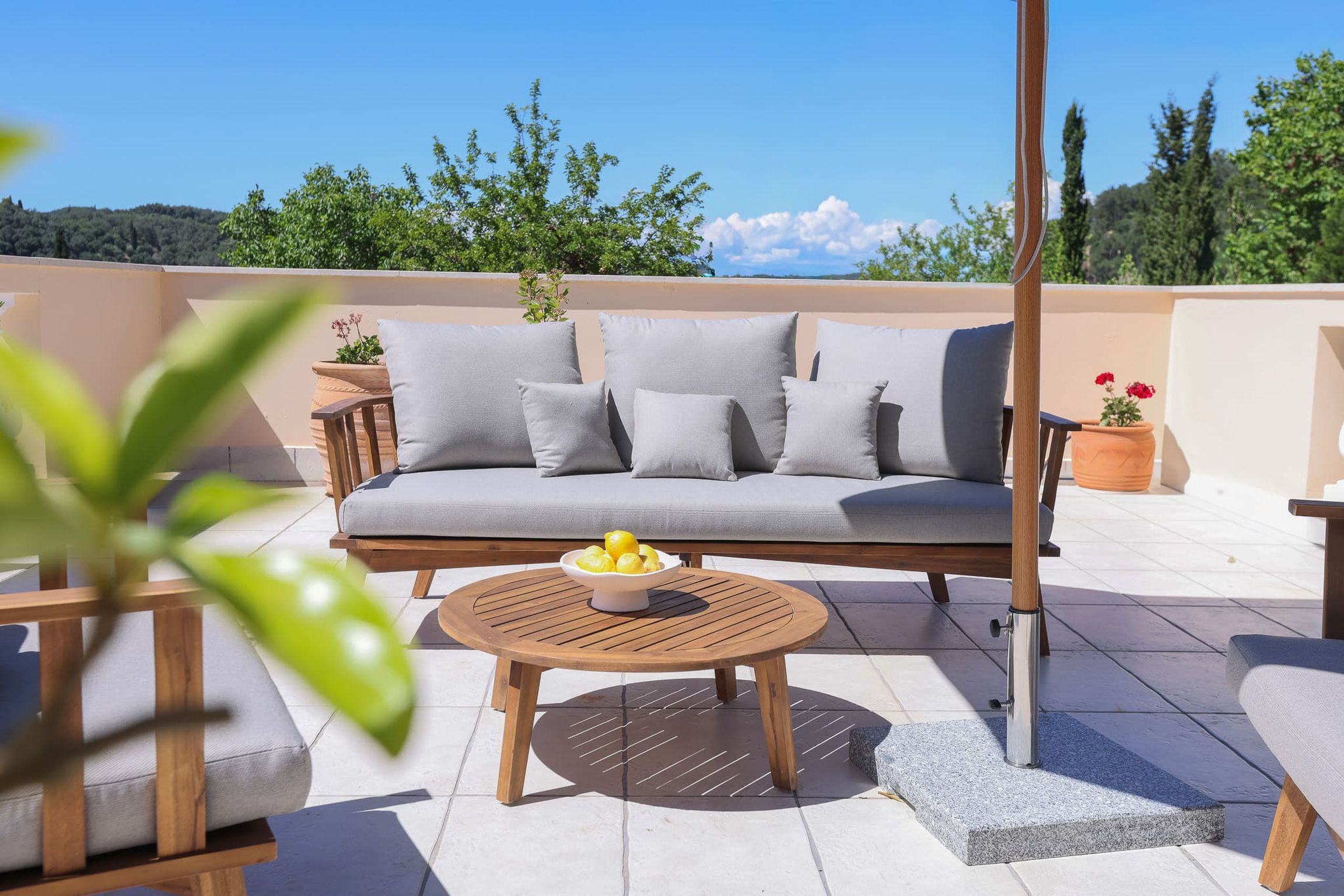 A sunlit outdoor seating area with a sofa, cushions, a round wooden table with a bowl of lemons, and potted plants.