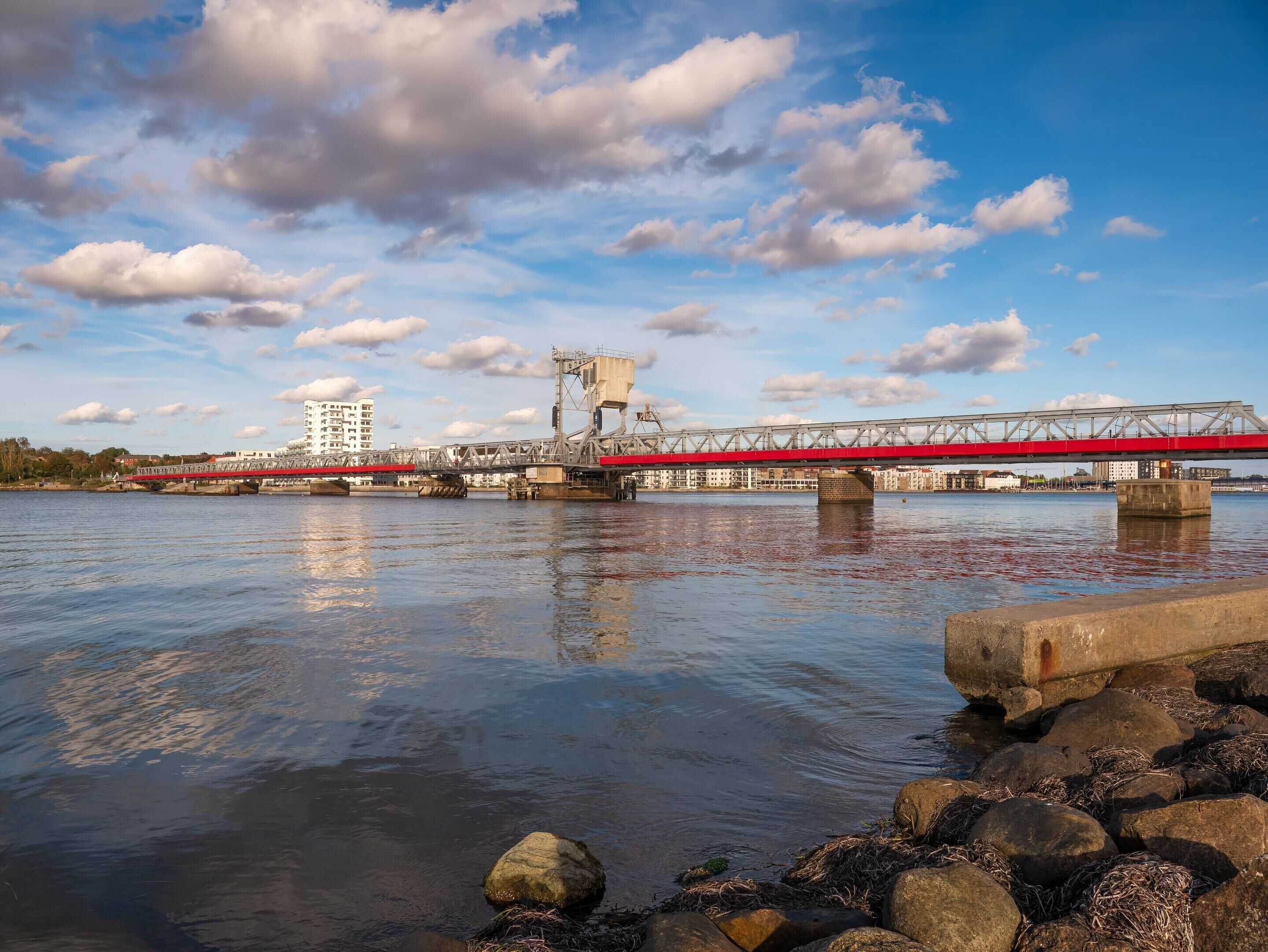 Un río con un puente, orilla rocosa y edificios al fondo.