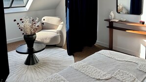 Una habitación con tragaluz, una silla blanca, una mesa redonda con un jarrón, un espejo y una cortina oscura.