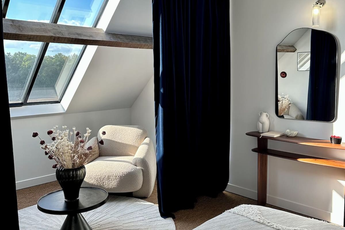 A room with a skylight, a white chair, a round table with a vase, a mirror, and a dark curtain.