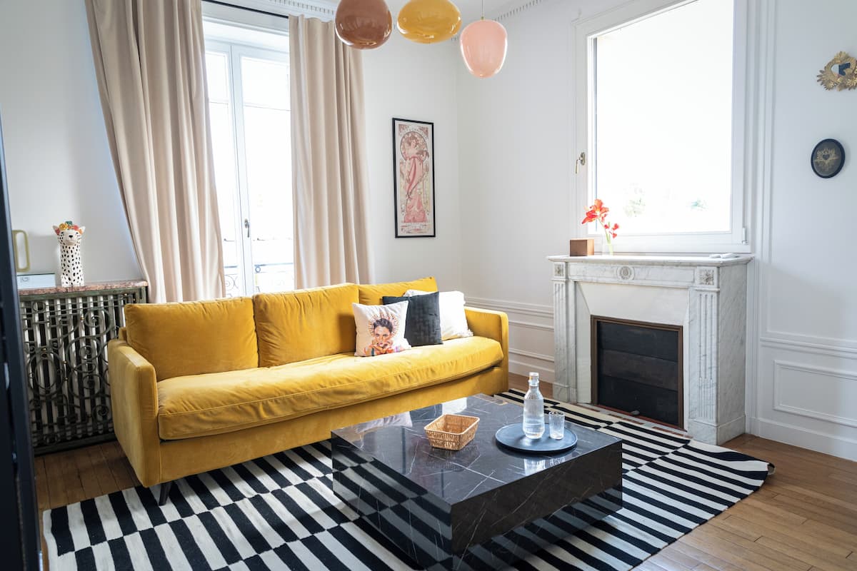 A living room with a yellow sofa, a black and white striped rug, a fireplace, and a large window with curtains.