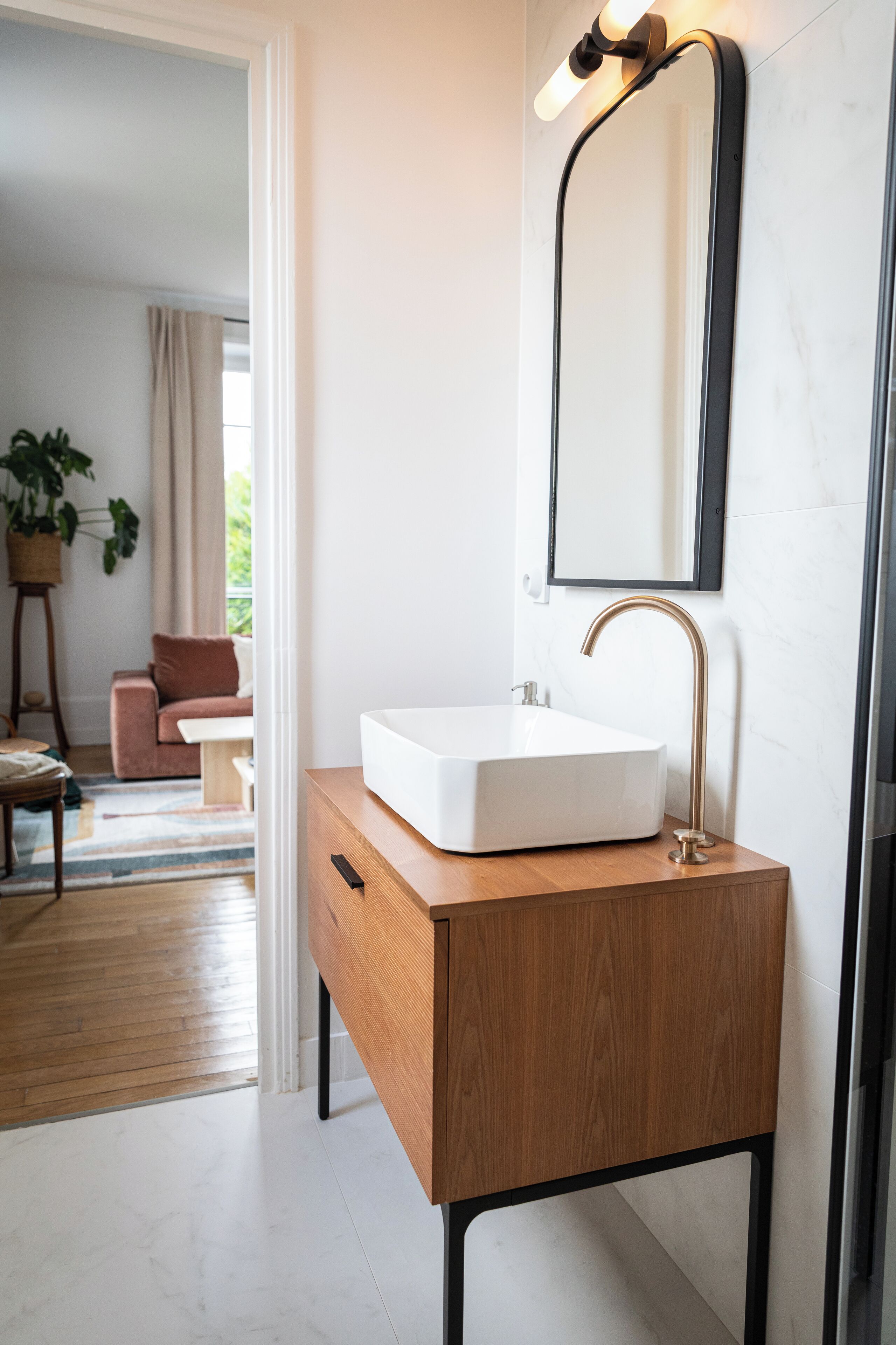A bathroom with a white sink, a wooden vanity, a black-framed mirror, and a potted plant.