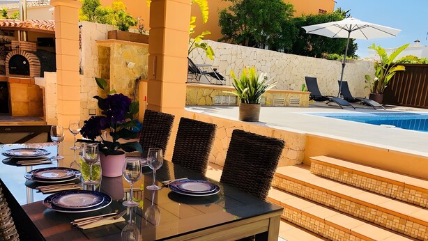 An outdoor dining area with a table set for four, surrounded by wicker chairs, near a swimming pool. - Villas 2 meet Estamarell – Private Oasis in Mallorca’s Natural Landscape (Romàntica)