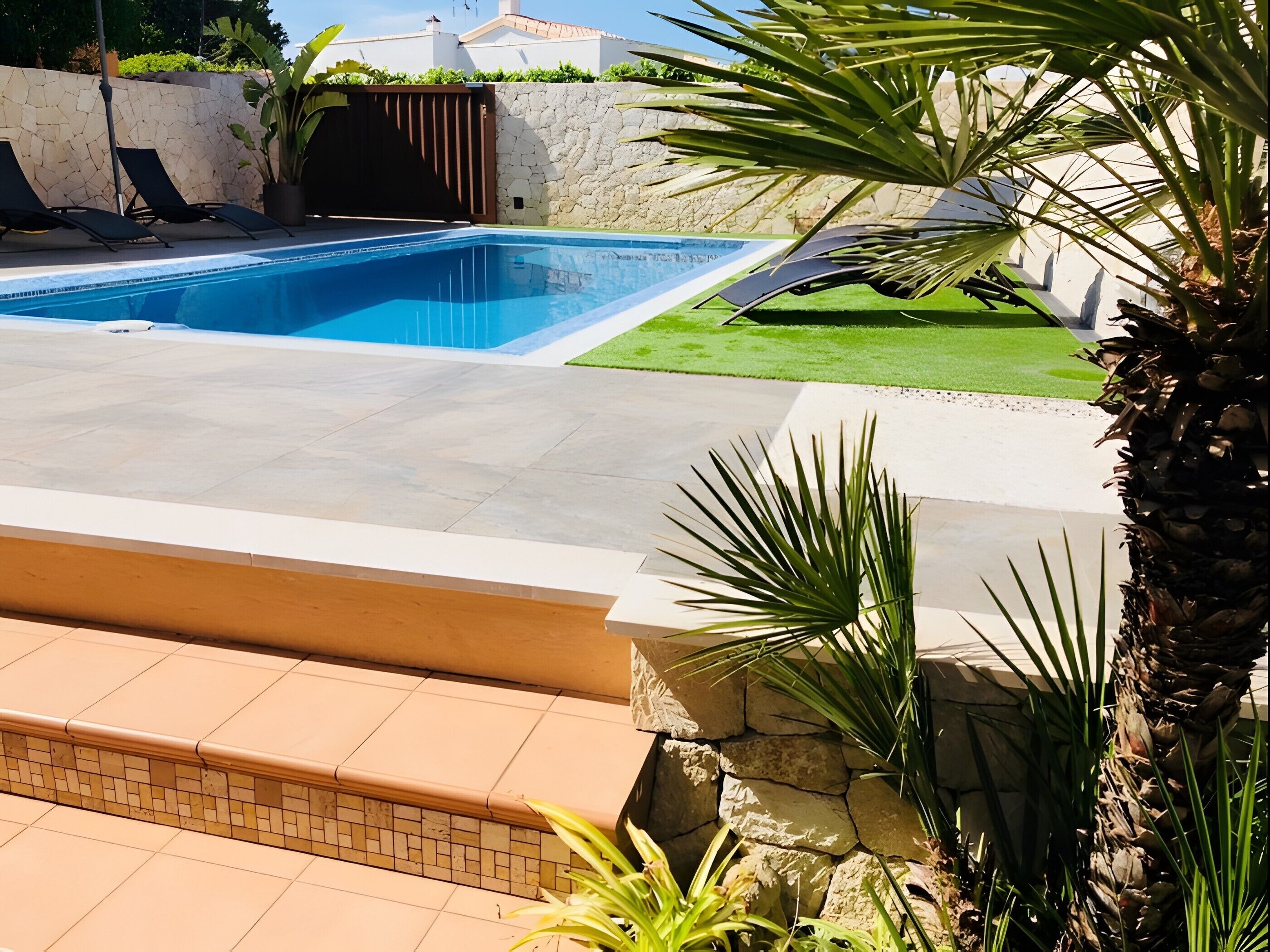 Una piscina con un muro de piedra, césped y una palmera.