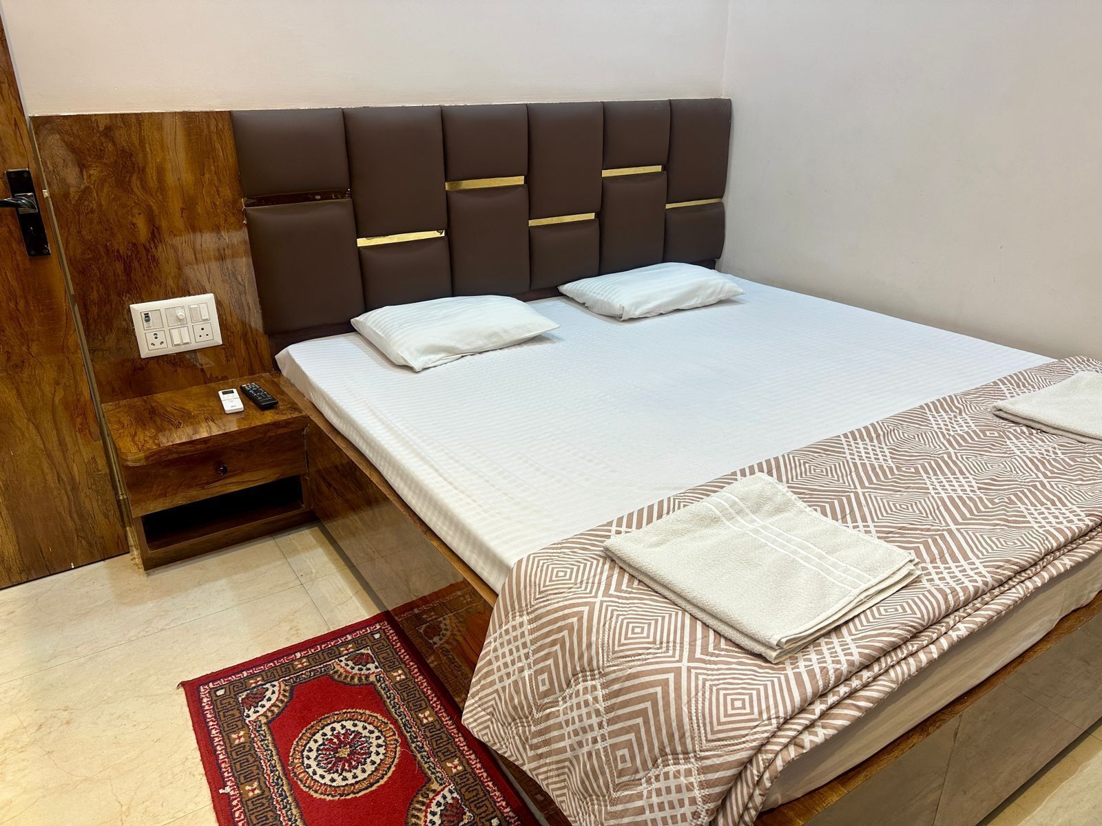 A hotel room with a bed, a bedside table, a power outlet, and a red patterned rug.