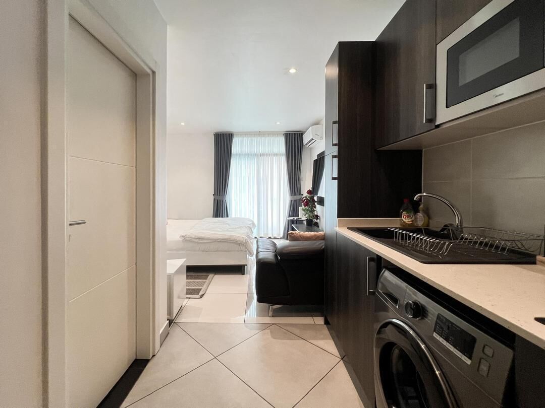 A compact hotel room with a kitchenette, a washing machine, and a bed with a view of the city.