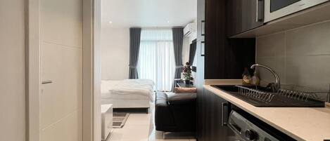 A compact hotel room with a kitchenette, a washing machine, and a bed with a view of the city.