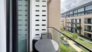 A balcony with a glass railing, a round table, and two chairs. The balcony overlooks a courtyard with greenery and a modern building in the background. - Embassy Gardens (Accra)