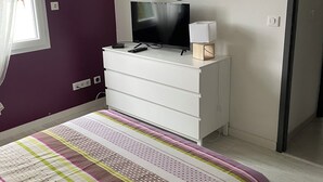 Une chambre à coucher avec un lit, un téléviseur posé sur une commode, un miroir et une fenêtre avec des rideaux.