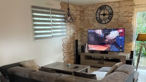 Un salon moderne avec un canapé d’angle, un téléviseur à écran plat fixé à un mur en pierre, un luminaire au plafond et une fenêtre équipée de stores.