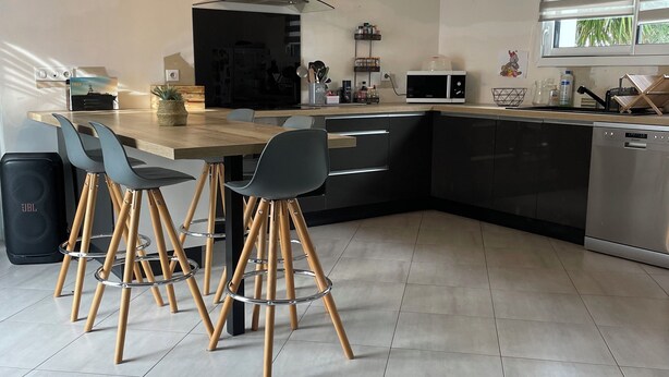 Une cuisine moderne dotée d’un îlot central, de meubles noirs et d’un four à micro-ondes. On y trouve trois chaises avec des pieds en bois et un coussin noir.