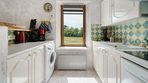 A compact kitchen with a window, a washing machine, and a coffee maker. - Holiday Home "Gîte Les Hortensias with Pool" – Private Pool, Wi-Fi & Air Conditioning (Trébédan)