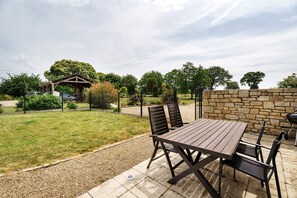 Een terras buiten met een houten tafel en stoelen, een stenen muur en een pergola.