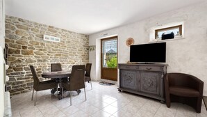 A room with a stone wall, a dining table with chairs, a TV on a cabinet, and a window with a view. - Holiday Home "Gîte Les Hortensias with Pool" – Private Pool, Wi-Fi & Air Conditioning (Trébédan)