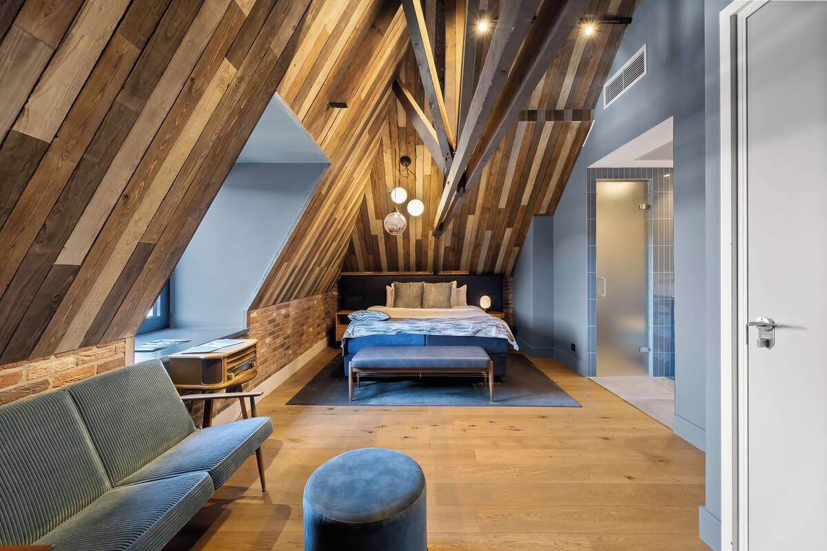 A modern loft bedroom with exposed wooden beams, a large bed, a seating area, and a small table with a vintage radio.