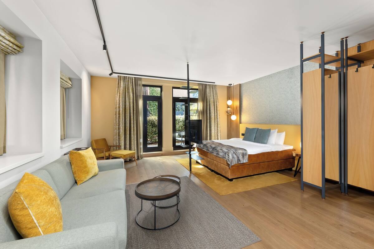 A modern hotel room with a large bed, a sofa, a small round table, and a view of the outdoors through large windows.