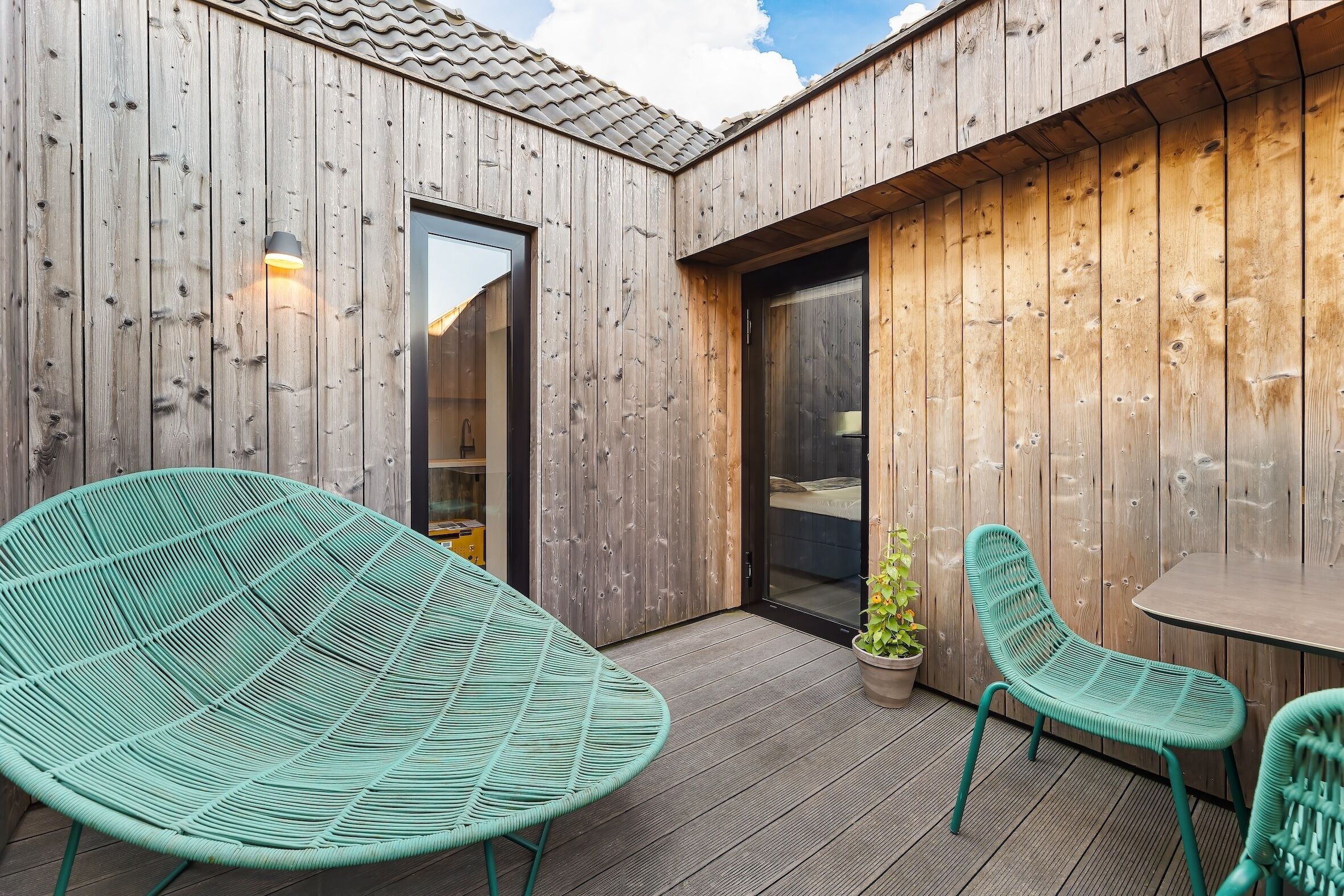 Una terraza de madera con dos sillas verdes y una mesa, una puerta que da a una habitación y una planta en maceta.