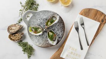 A dish of oysters on ice with herbs, accompanied by lemon halves and a wooden board with a napkin and forks.