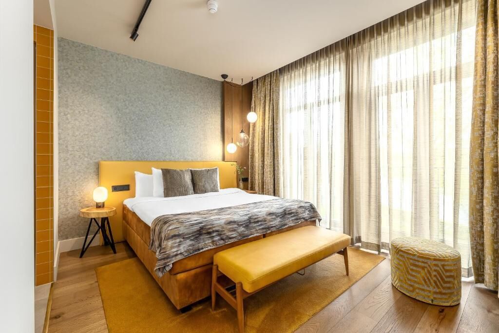A modern hotel room with a large bed, bedside lamps, a bench, and a view of the outdoors through sheer curtains.