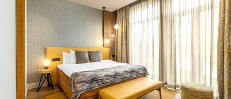 A modern hotel room with a large bed, bedside lamps, a bench, and a view of the outdoors through sheer curtains.