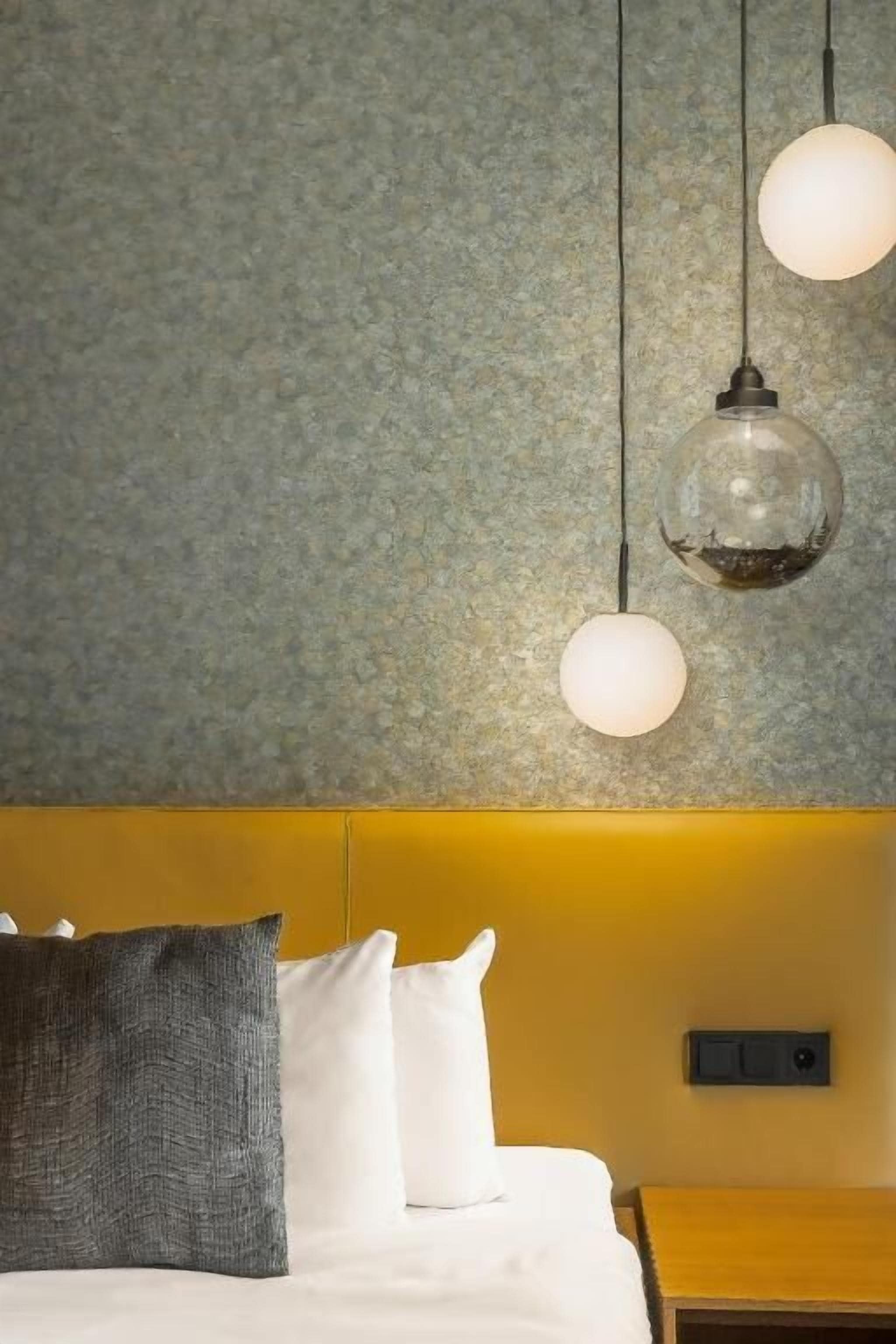 A hotel room with a textured gray wall, a yellow headboard, and a bed with white and gray bedding. There are three hanging spherical light fixtures.