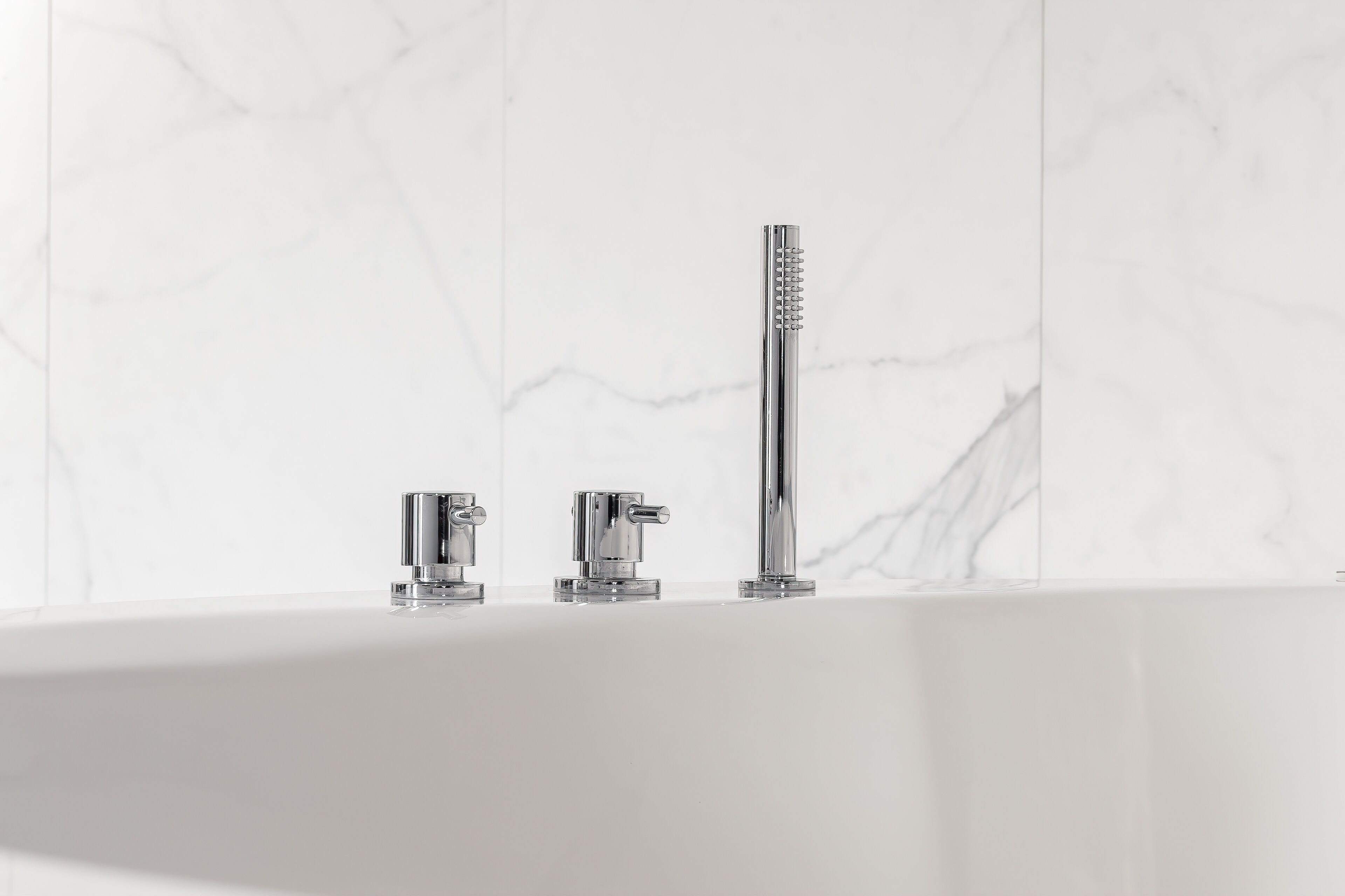 Un baño con pared de mármol blanco, un lavamanos blanco y tres grifos de cromo.