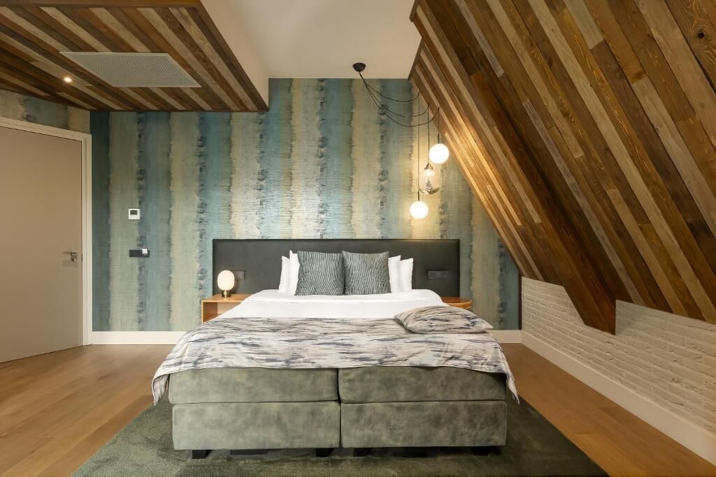 Un dormitorio con techo inclinado, cama con cabecero tapizado en gris, mesita de noche adosada a la pared y pared con diseño azul texturizado.