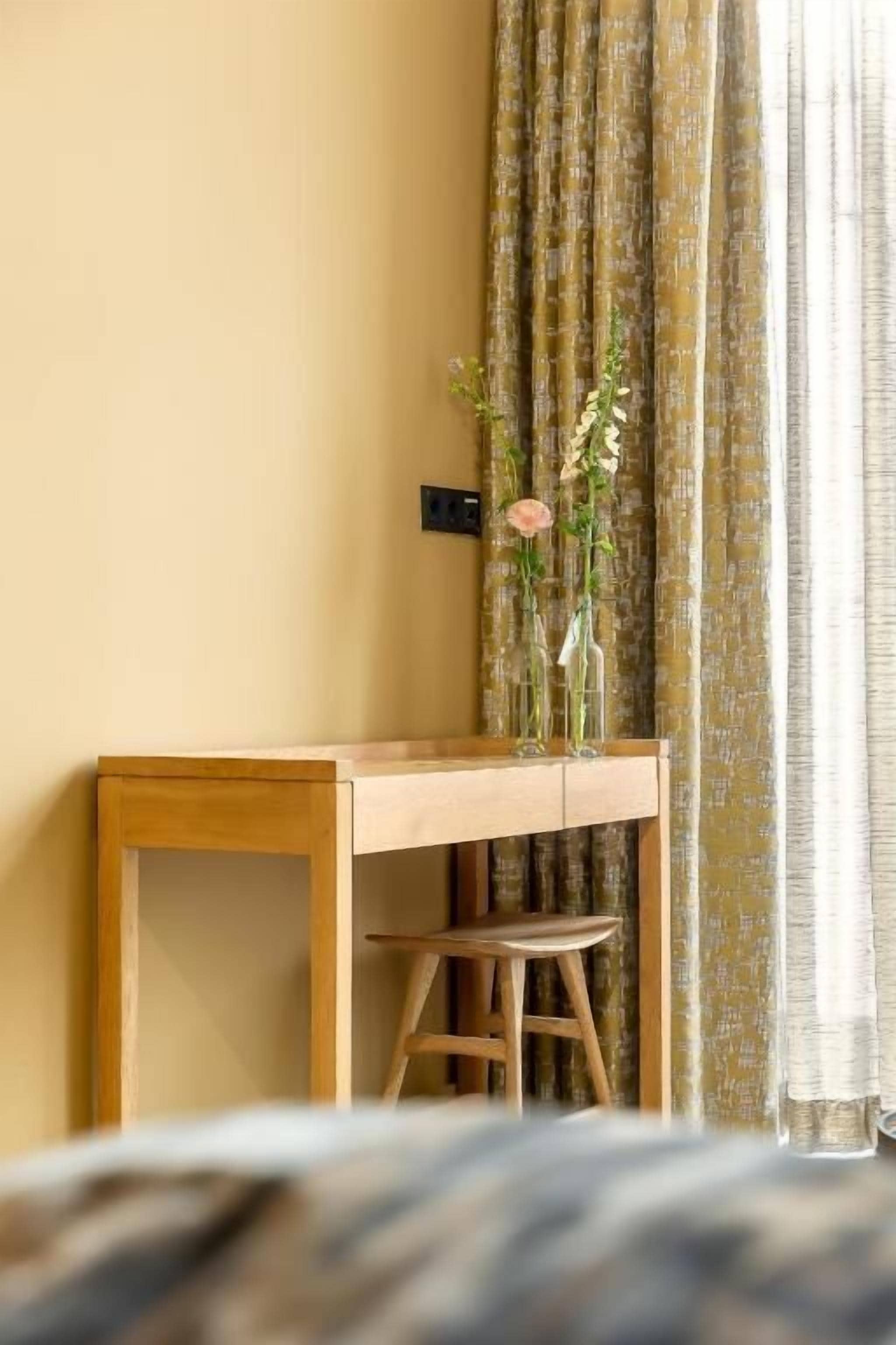 Una habitación con un escritorio de madera, una silla, un florero con flores y cortinas.