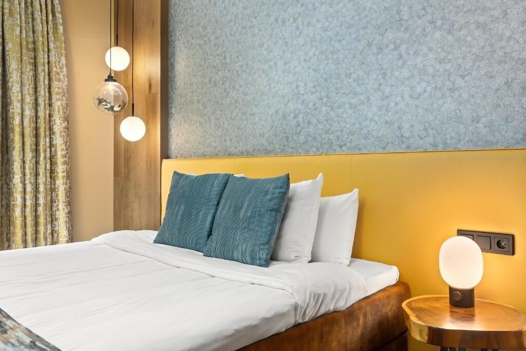 Ein ordentlich bezogenes Bett mit weißer Bettwäsche und türkisfarbenen Kissen, ein Holz-Kopfschild und ein runder Nachttisch mit einer Lampe.
